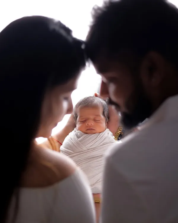 An artistic silhouette shot focusing on the peacefully sleeping newborn. The parents frame their baby, their heads touching in a moment of quiet reverence and love. The backlighting creates a soft, ethereal glow.