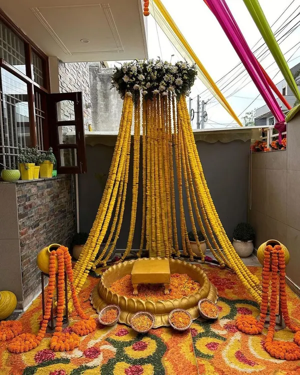 A traditional Haldi setup in a home's courtyard, featuring a canopy of marigold strings, a brass urli for the ceremony, and a floral rangoli on the floor.