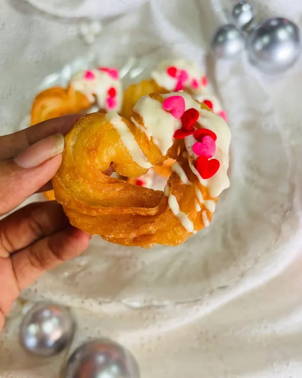Here is a close-up of my signature heart-shaped churro. You can see the crispy, golden texture of the churro, generously drizzled with creamy white chocolate and topped with red and pink heart-shaped sprinkles for a festive touch.
