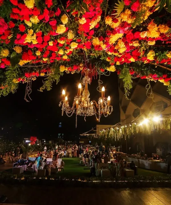 A breathtaking view of a rooftop wedding at night, with a massive floral arch of red and yellow flowers illuminated by a grand chandelier.