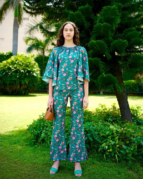 A full-length shot from a summer collection, featuring a green floral co-ord set. The outdoor garden setting and natural light create a fresh, vibrant look perfect for resort wear.