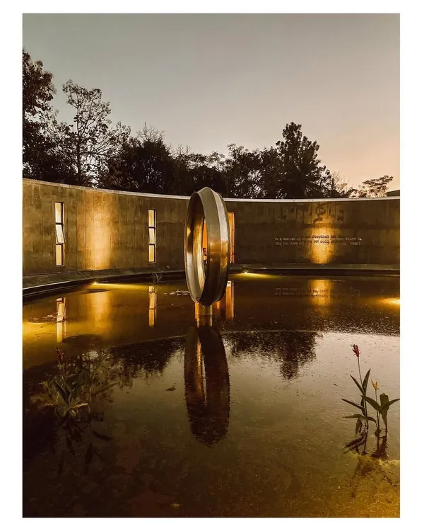 The Dr. Vishnuvardhan Memorial Complex at night, where strategic lighting highlights the texture of the concrete walls and the central ring sculpture. The perfect reflection in the water creates a sense of balance and peace, fulfilling the space's purpose as a place for remembrance. This image also covers media ID 11.