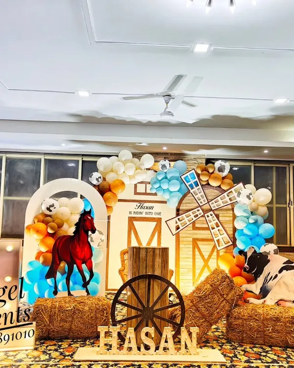 A fun cowboy and farm theme birthday decoration. This setup includes a barn-style backdrop, hay bales, a wooden wagon wheel, and character cutouts of a horse and cow, perfect for a rustic party.