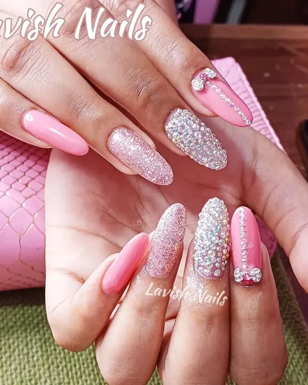 A glamorous set of long, almond-shaped acrylic nails. This design features a mix of solid pink, sparkling pink glitter, and accent nails fully encrusted with rhinestones, topped with a delicate 3D bow for a truly statement look.