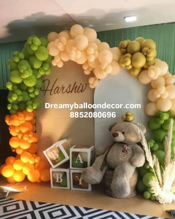 This is a unique jungle-themed decoration I did for a baby celebration. It features a balloon arch in sage green, orange, and beige, with a large teddy bear, B-A-B-Y boxes, and a custom name sign for "Harshiv".