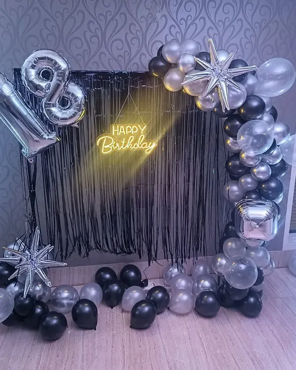 A sleek and modern setup for an 18th birthday. This decoration features a black fringe curtain, a half-arch of black, silver, and clear balloons, starburst foils, and a 'Happy Birthday' neon sign.