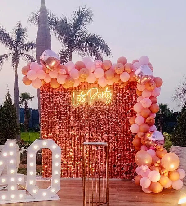 A chic outdoor 40th birthday celebration. This setup features a rose gold sequin wall, a "Let's Party" neon sign, and large marquee numbers on a wooden deck, perfect for an evening party.