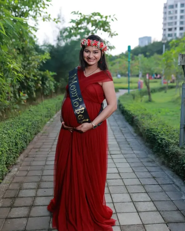 A happy mom-to-be in a simple yet elegant red maternity gown, perfect for an outdoor photoshoot. The empire waistline and soft tulle are designed for a comfortable fit while looking beautiful.