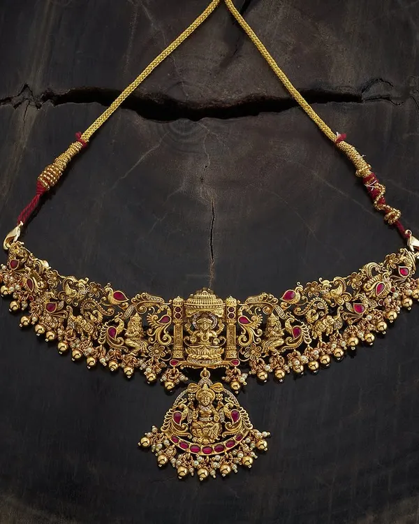An antique gold-toned temple choker necklace featuring a central pendant of Goddess Lakshmi. The entire piece is detailed with peacock motifs and ruby-colored stones, embodying sacred design and traditional artistry.