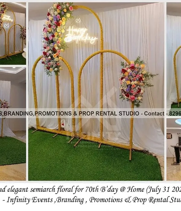 A simple and elegant floral setup for a 70th birthday celebration at home. The decor features golden semi-arches adorned with beautiful flowers and a "Happy Birthday" neon sign.
