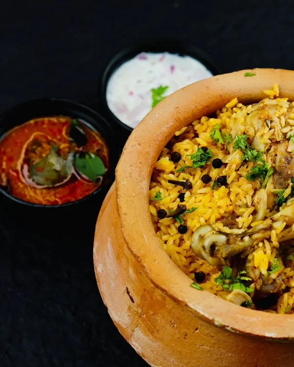 A serving of our wood-fired chicken biryani in a clay pot, accompanied by bowls of tangy Baigan ka Khatta and creamy raita. This is the complete meal experience we deliver for your events.