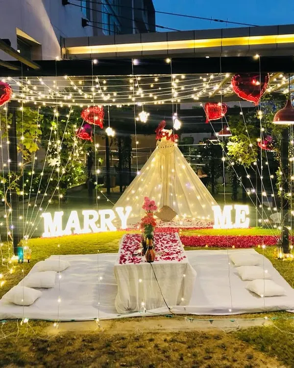 A complete garden proposal package featuring a fairy light cabana for a romantic dinner and a separate teepee tent area. The scene is set with large 'MARRY ME' marquee letters and a path of red rose petals.