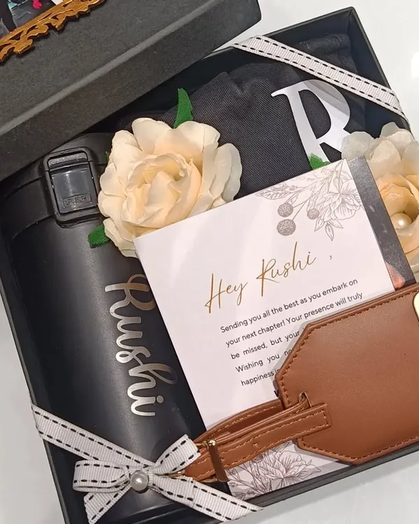 A farewell gift box curated to wish someone the best for their next chapter. This hamper includes a coffee sipper and a leather luggage tag, both personalized with the name 'Rushi', along with other goodies in a classic black box.
