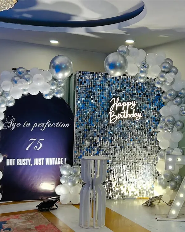 A sophisticated 75th birthday setup featuring a silver shimmer wall paired with a white "Happy Birthday" neon sign. The combination creates a modern and elegant look.