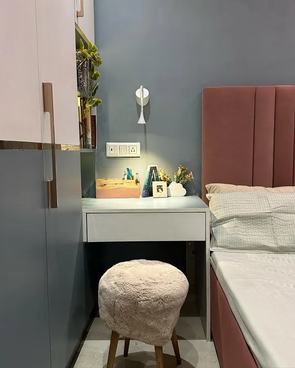 This guest room corner shows a compact and functional design. A small vanity desk is neatly tucked beside the bed, paired with a textured stool. The two-tone wardrobe with rose gold handles adds a touch of modern elegance, while the playful wall paint detail brings character to the space.