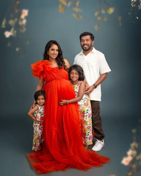 A beautiful family portrait celebrating their growing family. The mother's vibrant red gown stands out against the classic studio backdrop, while her two daughters lovingly embrace her baby bump.