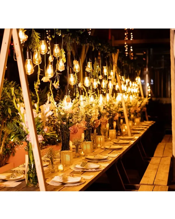 The full view of our "Blooms & Barrels" anniversary dinner setup. The combination of rustic wooden tables, hanging lights, and abundant fresh flowers creates a warm and inviting atmosphere for a memorable evening.