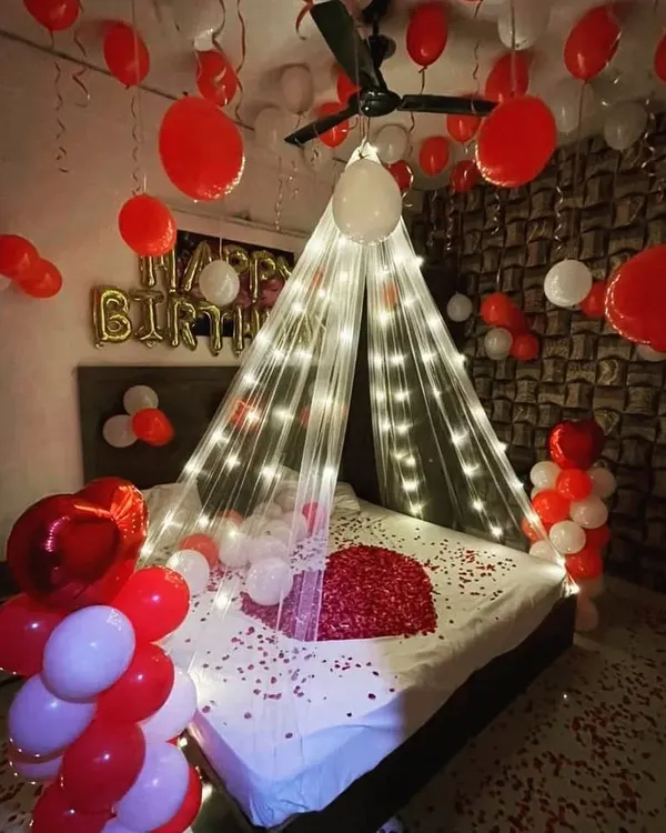 A classic romantic bedroom decoration, perfect for birthdays or anniversaries. This popular design features a sheer canopy with fairy lights over the bed, a heart made of rose petals, and dozens of red and white balloons floating on the ceiling.