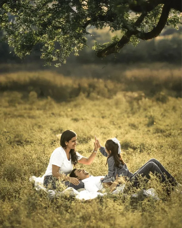 A family enjoys a playful moment during a picnic in a sunlit meadow. The father lies back while the mother and daughter share a high-five, their laughter echoing in the warm, golden light. This is a perfect example of a candid family photoshoot.