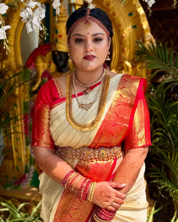 A frontal portrait of this beautiful bride, ready for her muhurtham. The makeup features a classic combination of defined eyes and a deep red lip, designed to harmonize with her traditional cream and red silk saree and gold jewellery.