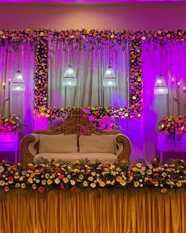 A luxurious wedding stage featuring a floral-framed backdrop, elegant hanging lamps, and dramatic purple lighting. This design creates a grand and romantic setting for the main ceremony.
