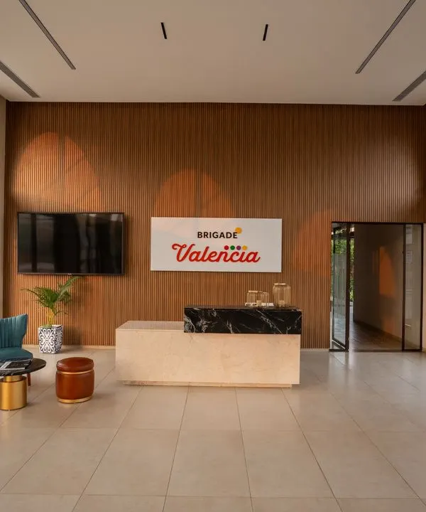 The reception of the Brigade Valencia marketing office features a double-height ceiling and large arched windows. We used a warm wood-paneled wall and a sleek marble-topped desk to create a welcoming first impression.