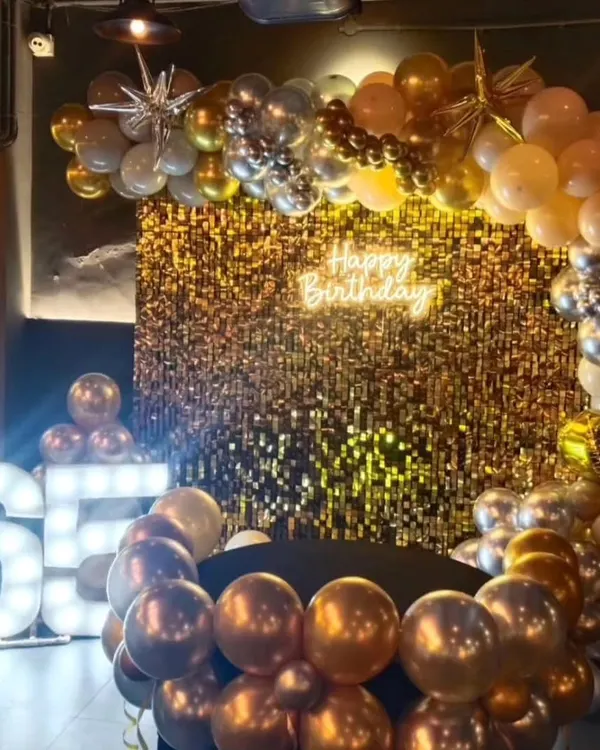A glamorous setup for a 65th birthday celebration. The decor features a stunning gold sequin wall, a balloon arch in chrome and metallic tones, a 'Happy Birthday' neon sign, and large light up marquee numbers.