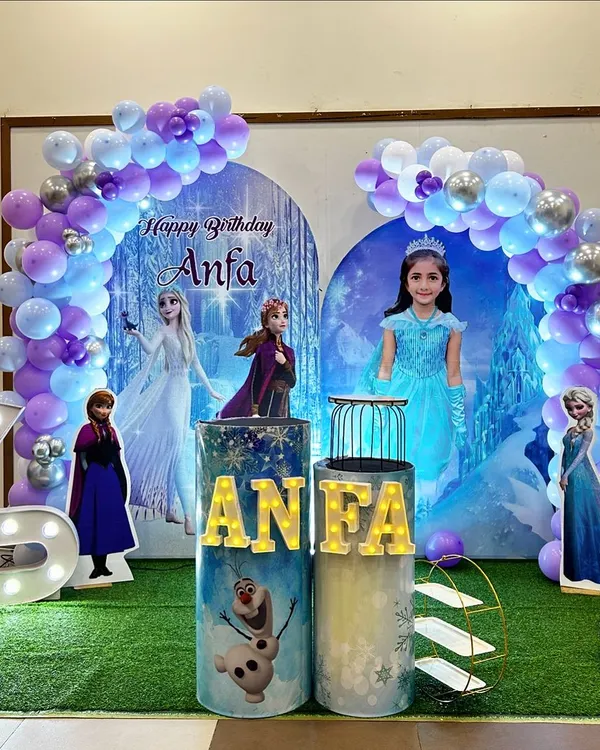 A complete Frozen-themed birthday party setup for Anfa's 6th birthday. It features character cutouts of Elsa, Anna, and Olaf, custom-printed pedestals, a light-up marquee number '6', and a balloon arch in shades of purple, blue, and silver.