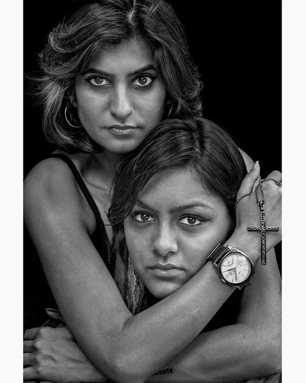 An intense black and white portrait of two women, focusing on their direct gaze and protective embrace. The high-contrast lighting emphasizes the raw emotion and connection between the subjects.