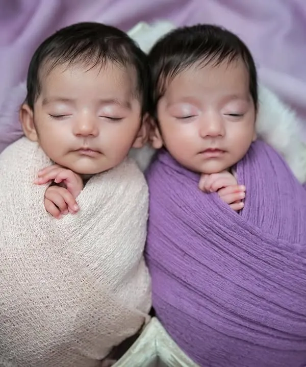 A close-up of newborn twins nestled together in a heart-shaped prop. Swaddled in complementary shades of pink and purple, this image beautifully highlights their peaceful bond.
