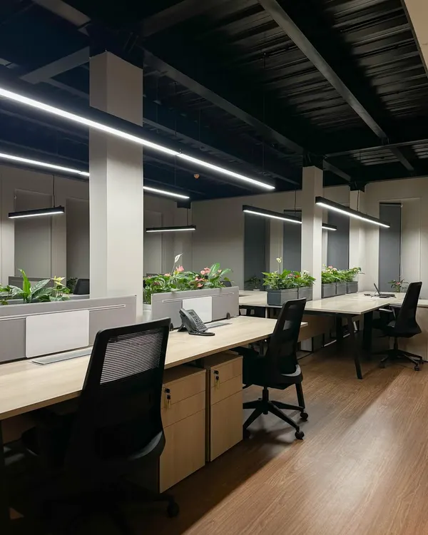 A view across the open-plan work floor, featuring rows of workstations with integrated planters. The industrial-inspired exposed ceiling is fitted with modern linear lighting to ensure a bright and energizing environment.