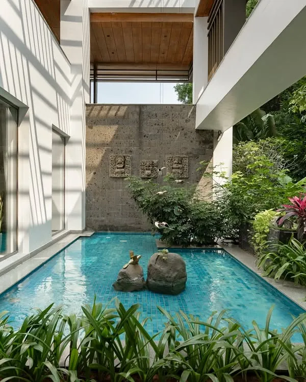 A tranquil pool nestled between the wings of a modern house, surrounded by lush tropical plants with red and green foliage. The design creates a private, resort-like escape right at home.