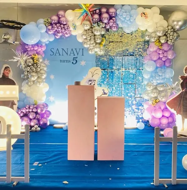 A magical Frozen theme for a 5th birthday. The backdrop features a shimmering blue waterfall effect, surrounded by a balloon arch in shades of purple, blue, and silver, with cutouts of Anna and Elsa.