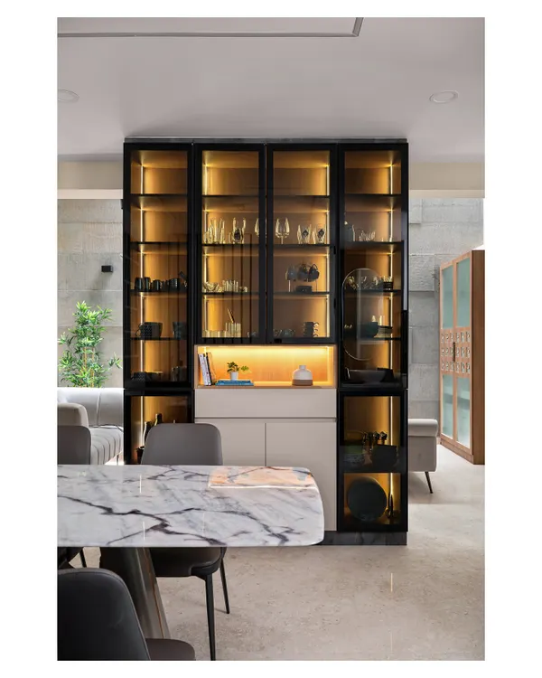 This stunning setup features a sleek, black-framed display unit with warm internal lighting, perfect for showcasing crockery and decor. The unit is paired with a beautiful marble-top dining table for a touch of class.