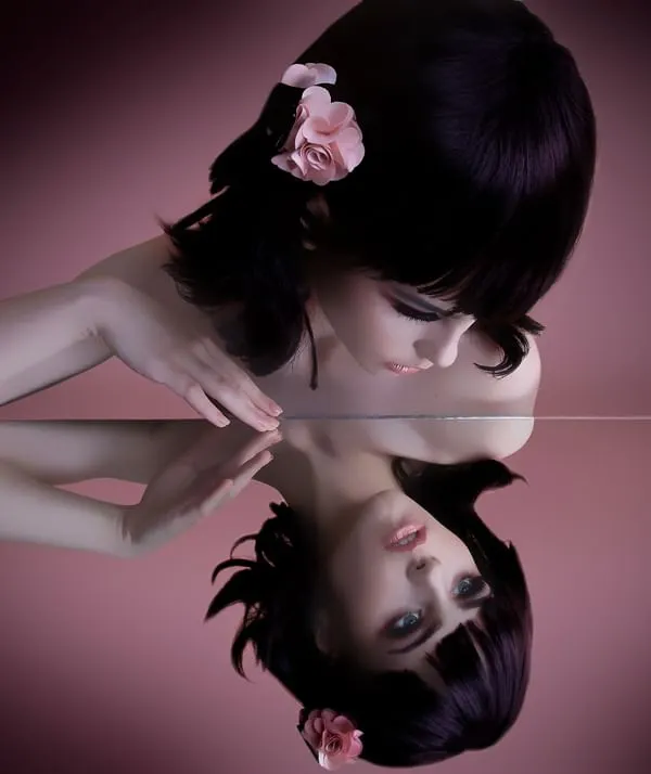 A creative beauty portrait showing a model with a sleek black bob and a pink flower in her hair, gazing at her own reflection in a mirror. This image showcases my technical skill with lighting and composition for artistic and conceptual photoshoots.