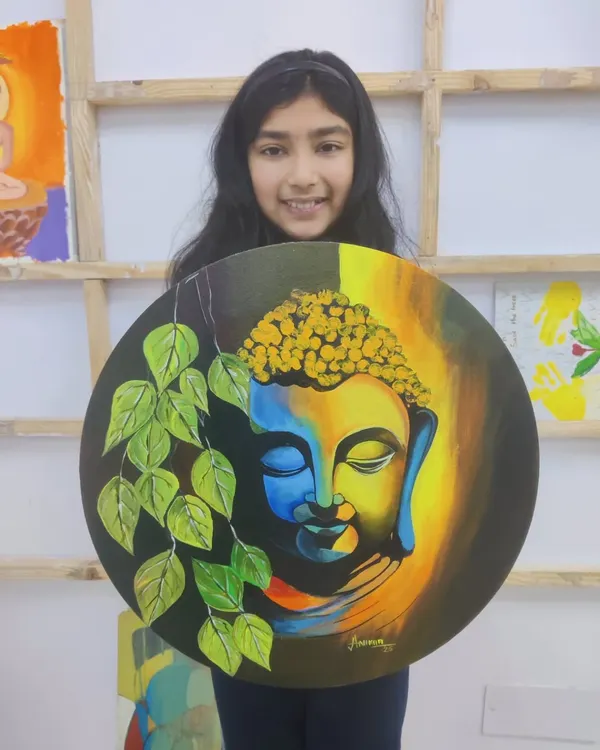 A young student proudly holds her beautifully finished acrylic painting of Buddha on a round canvas, a piece that radiates peace and serenity.