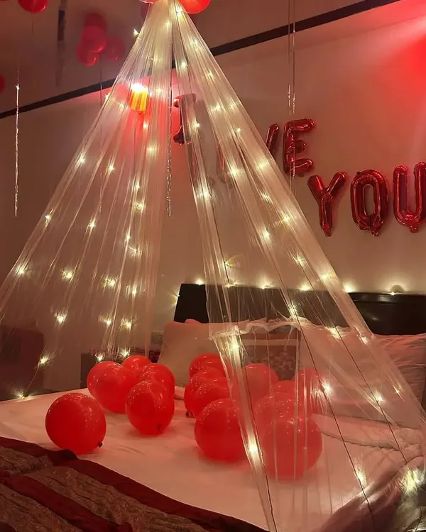 A beautiful and intimate proposal or anniversary setup. This design features a fairy light canopy over the bed, red heart-shaped balloons, and "I Love You" foil letters in the background.