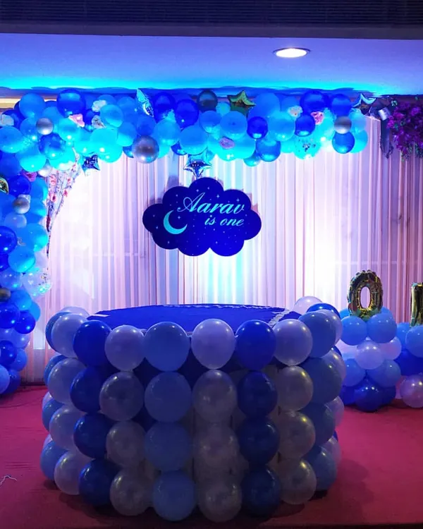 For this first birthday party, I created a complete 'moon and stars' themed stage. The setup includes a custom "Aarav is one" name sign, a balloon-wrapped cake table, and a full balloon arch, all in a classic blue and white color scheme for a baby boy.