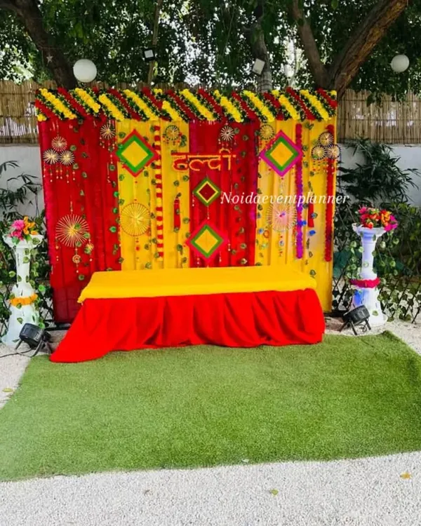 A classic Haldi ceremony setup with a bright yellow and red backdrop, decorated with marigold flowers and traditional hangings for a festive feel.