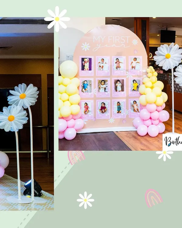 A sweet and cheerful 'My First Year' photo board with a daisy theme. The arched display is decorated with pastel pink and yellow balloons and features large, charming daisy props on either side.