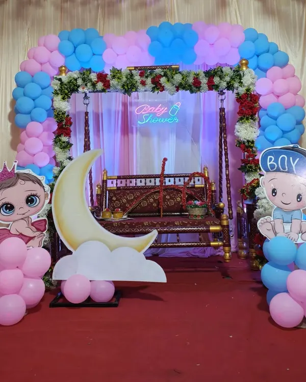 A beautiful setup for a traditional baby shower or Dohale Jevan. It features a decorated swing (jhula), a pink and blue balloon arch, and cute baby cutouts to welcome the little one.