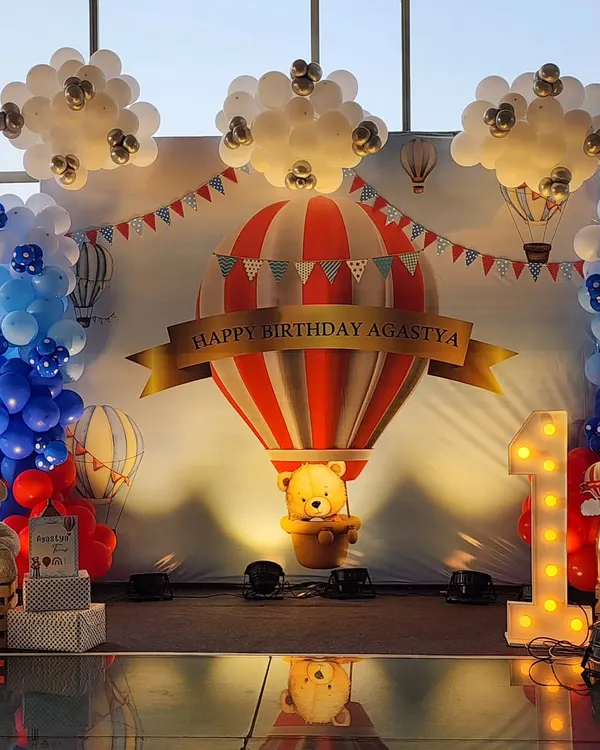 A grand first birthday celebration with a hot air balloon theme. This custom backdrop features a large balloon with a teddy bear, surrounded by balloon clouds and a lit-up "ONE" marquee sign, creating a perfect photo opportunity.