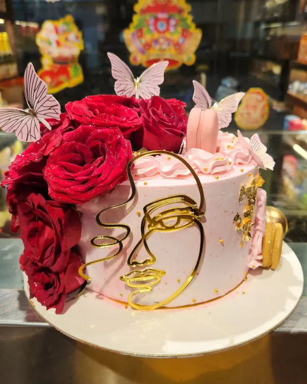 An artistic and elegant cake perfect for a sophisticated birthday or celebration. This design features a delicate pink frosting, a cascade of fresh red roses, edible gold leaf, and a modern gold wire-art face topper for a truly stunning centerpiece.
