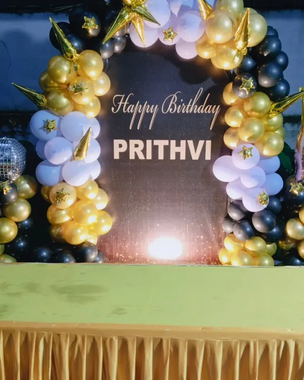 A stylish black, gold, and white balloon arch for Prithvi's birthday, complete with a shimmer backdrop, star-shaped balloons, and a champagne bottle prop.