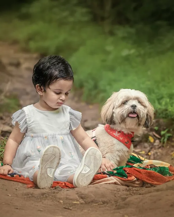 A little girl sits on a blanket on a forest path next to her fluffy white dog. This adorable portrait captures the sweet and innocent friendship between a child and her furry companion.