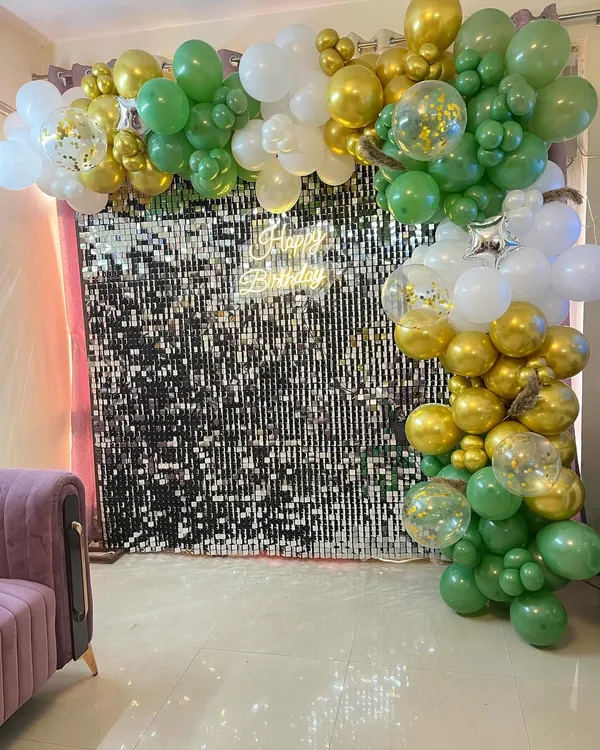 A stylish birthday backdrop featuring a shimmering silver sequin wall. It is framed by a green, gold, and white balloon arch and includes a 'Happy Birthday' neon sign.