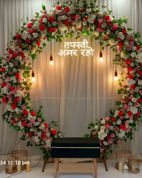 A full circular arch made of red and white flowers for a Tapasvi ceremony at home. The neon sign and hanging Edison bulbs add a modern touch to the traditional setup.