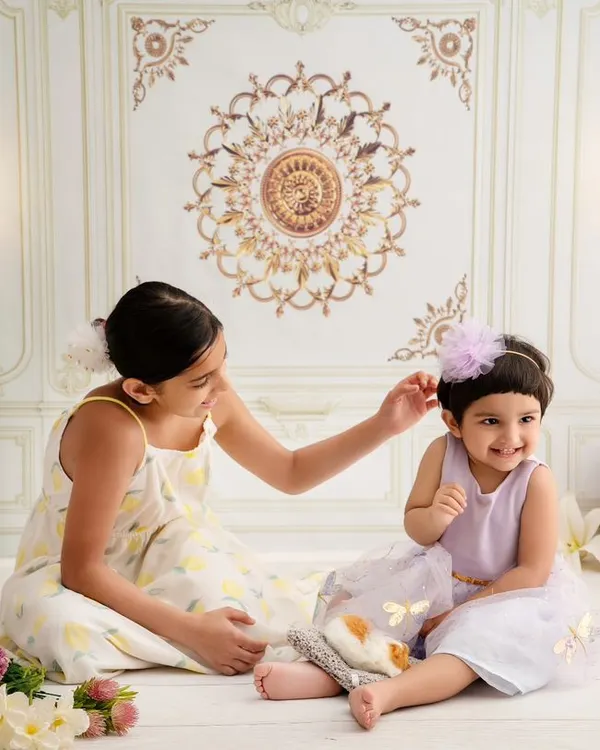 A sweet, candid moment between two sisters. The older sister gently adjusts the younger one's hair bow in our elegant, light-filled studio, showing a quiet moment of care.
