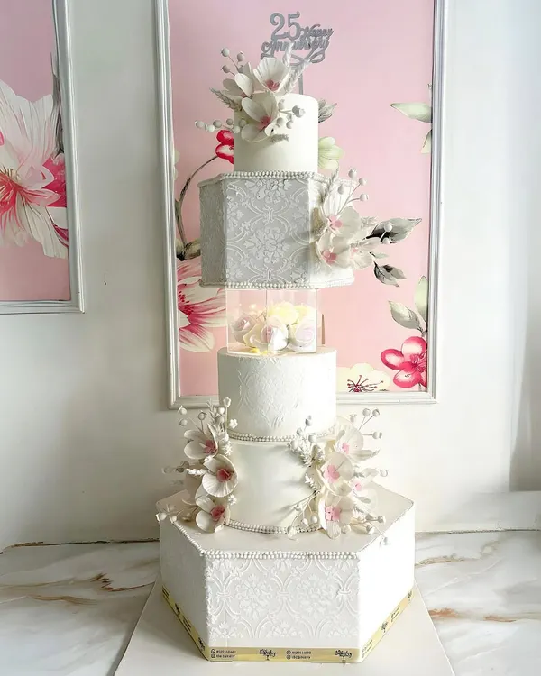 A grand creation for life’s grandest moments. This five-tiered white anniversary cake is a showstopper, featuring hexagonal and square tiers, intricate textures, and delicate, handcrafted sugar flowers.
