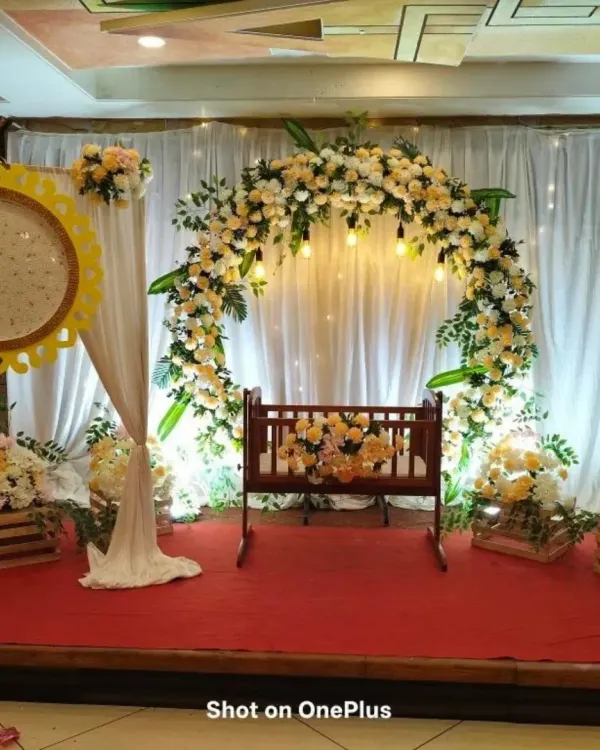 A full view of a naming ceremony stage, showing the decorated cradle under a soft floral arch. The setup includes matching flower arrangements in rustic wooden crates, creating a cohesive and charming look.